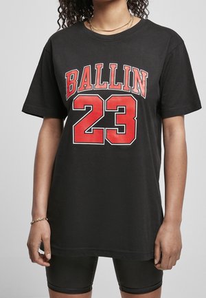 Persoon draagt een zwart oversized t-shirt met rode tekst "BALLIN 23" en zwarte aansluitende shorts, gecombineerd met een gouden ketting en armband.