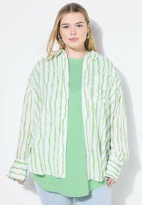 Camicia a righe verdi con bottoni, indossata sopra una maglietta verde unita, abbinata a jeans in denim chiaro con dettagli in perle. Tessuto morbido e vestibilità rilassata.