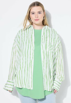 Camicia a righe verdi con bottoni, indossata sopra una maglietta verde unita, abbinata a jeans in denim chiaro con dettagli in perle. Tessuto morbido e vestibilità rilassata.