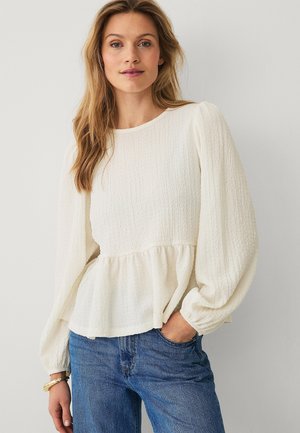 Kvinde med langt blondt hår iført en hvid bluse med tekstur og puffede ærmer samt blå jeans, stående med den ene hånd i lommen.