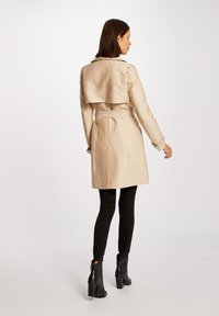 Beige trenchcoat med bältad midja, slagkrage och ryggdetalj i kapor, kombinerad med svarta slim-fit byxor och ankelskor.