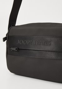 Borsa a tracolla nera realizzata in tessuto morbido, con una tracolla nera testurizzata e il logo inciso "JOOP! JEANS" lungo una tasca con zip.