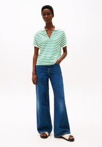 Striped grønn og hvit polo skjorte med v-hals og korte ermer, sammen med høytlivs, vide blåjeans og hvite sandaler.