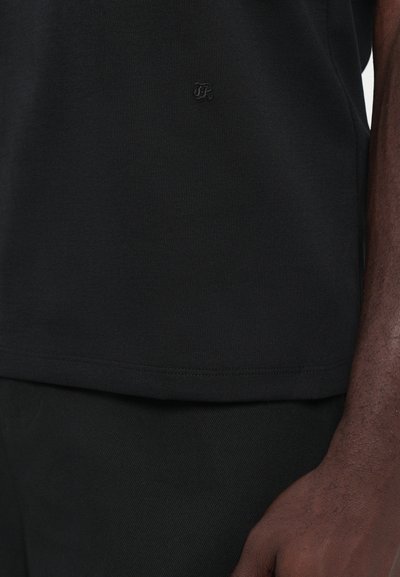 Schwarzes T-Shirt mit strukturiertem Stoff, Rundhalsausschnitt und einem kleinen, tonal gehaltenen Logo am Saum. Das Design ist einfach und unauffällig.