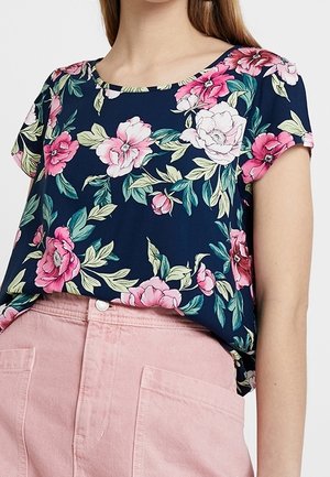 Femme portant un haut à manches courtes bleu marine avec un imprimé floral rose et blanc, rentré dans un pantalon taille haute rose clair avec des poches avant.