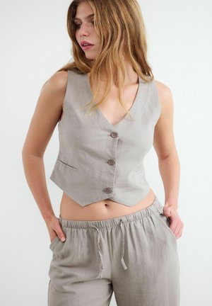 Vrouw draagt beige mouwloze, geknoopte vest en bijpassende losse broek met handen in de zakken, kijkt opzij.