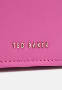 Superficie in pelle rosa con cuciture ordinate e lettere metallizzate dorate che formano la scritta "TED BAKER" centrata sopra la cucitura.