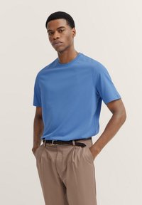 Blue short-sleeve shirt made of a smooth, lightweight fabric, paired with light brown trousers and a braided black belt.