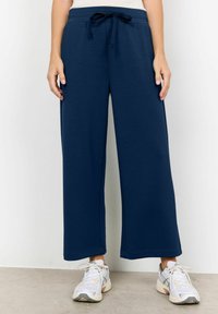 Navy blue wide-leg sweatpants with a drawstring waist, smooth texture, and contrasting stitching. Paired with light athletic shoes.