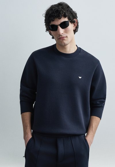 Sweat-shirt bleu marine à col rond et manches longues, avec un petit logo sur la poitrine. Texture lisse et coupe décontractée, assorti d'un pantalon coordonné.