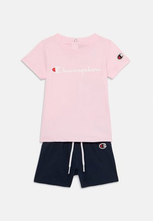 Camiseta de algodón rosa con el logo blanco de "Champion" y pantalones cortos azul marino con un parche de logo y cordón blanco. Conjunto casual de dos piezas.
