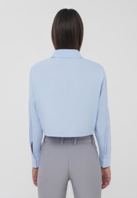 Camicetta azzurro chiaro a maniche lunghe con colletto, presenta polsini abbottonati e una texture liscia, abbinata a pantaloni grigi.