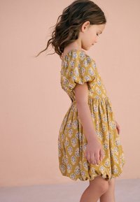 Mosterdgele jurk met pofmouwen, met een witte botanische patroon. Uitlopende zoom en een geplooide taille. Soepele stoffen textuur.