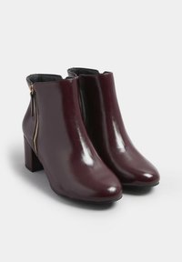 Par de botas de cuero burdeos oscuro hasta el tobillo con tacones de bloque y cremalleras laterales doradas sobre un fondo blanco.