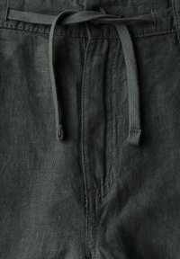 Tmavě zelené lněné kalhoty s elastickým pasem a hladkým povrchem, které mají viditelné stehy podél švů.