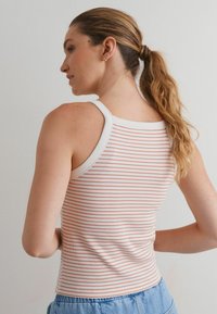 Femme avec une queue de cheval châtaine clair portant un débardeur sans manches blanc et rose rayé, ainsi que des pantalons en denim bleu clair, de profil.