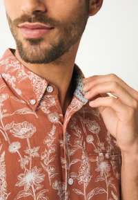 Chemise à boutons couleur rouille avec motifs floraux blancs, tissu texturé et col contrastant à motifs avec des accents bleus.