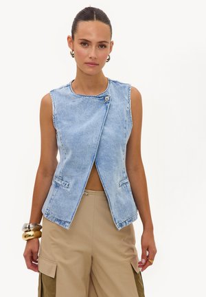 Femme aux cheveux coiffés en arrière porte un gilet en jean bleu clair sans manches avec des boutons sur le devant et un pantalon beige à jambes larges avec des poches cargo.