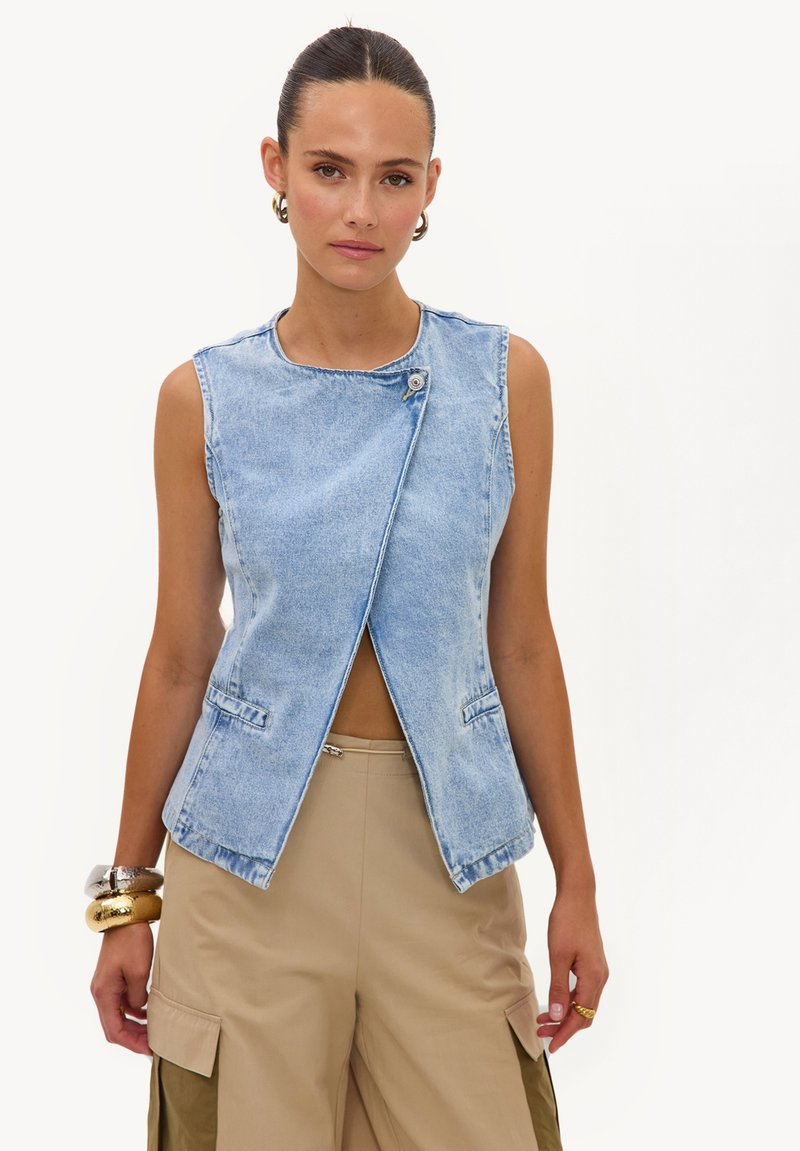 Femme aux cheveux coiffés en arrière porte un gilet en jean bleu clair sans manches avec des boutons sur le devant et un pantalon beige à jambes larges avec des poches cargo.