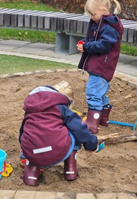 Två barn leker i en sandlåda. Den ena har på sig en vinröd och blå jacka med matchande gummistövlar; den andra har blå jeans och vinröda stövlar.