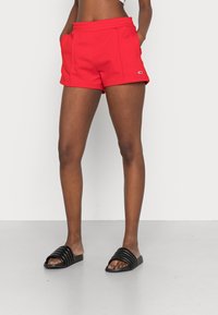 Shorts de sport rouges avec une taille élastique, deux poches latérales et un petit logo en accent. Assortis à des sandales noires à brides.