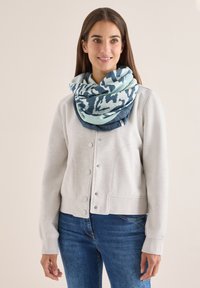 Lys grå sweatshirt med knaplukning foran, ledsaget af et stort blåt og teal mønstrede tørklæde viklet om halsen. Denim jeans er synlige.