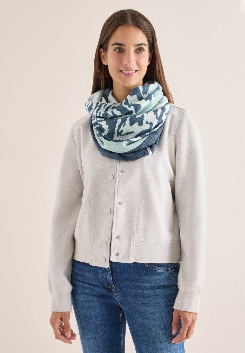 Lys grå sweatshirt med knaplukning foran, ledsaget af et stort blåt og teal mønstrede tørklæde viklet om halsen. Denim jeans er synlige.