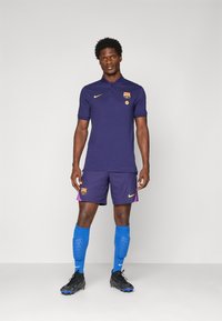 Barcelona jersey in dark purple with yellow accents, paired with purple shorts, blue socks, and black cleats. Features club logo and Nike branding.