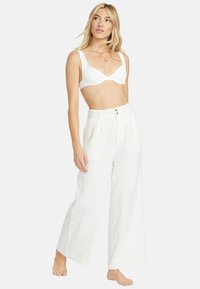 Vita linnebyxor med vida ben och hög midja, med två knappar, tillsammans med en vit strukturerad bh-top. Modellen står barfota.