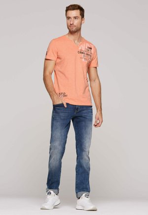 Mężczyzna stojący, ubrany w brzoskwiniowy t-shirt z grafiką, niebieskie jeansy z podwiniętymi nogawkami i białe trampki, patrzy lekko na bok.