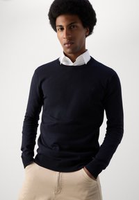 Navy blue sweater made of a smooth knit fabric, featuring a crew neck and ribbed cuffs, paired with a white collared shirt underneath.