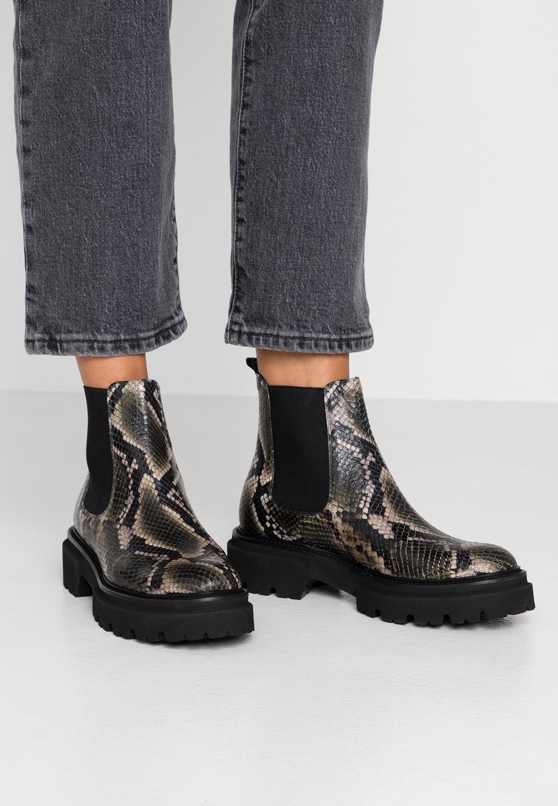 Botas de tobillo con suelas gruesas negras y patrón de serpiente, combinadas con jeans negros cortos, sobre un fondo liso.