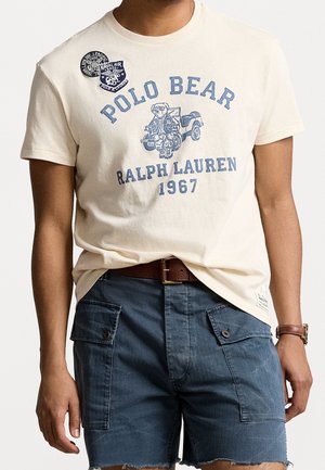 Mężczyzna ubrany w kremowy t-shirt Polo Bear Ralph Lauren 1967 z grafiką misia i jeepem, w połączeniu z niebieskimi szortami cargo i brązowym paskiem.