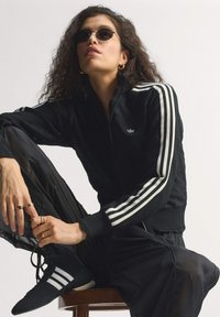 Woman in black Adidas track jacket, matching pants, and shoes with white stripes, wearing small black sunglasses and gold hoop earrings, seated.