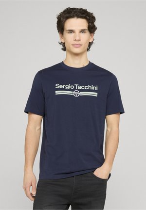 Junger Mann mit lockigem Haar, der ein dunkelblaues Sergio Tacchini T-Shirt und schwarze Hosen trägt, steht vor einem schlichten hellen Hintergrund.