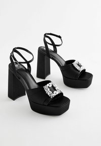 Black platform sandals featuring a satin finish, thick block heels, and a decorative jewel accent on the upper strap. Adjustable ankle straps included.