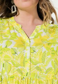Koszula z krótkim rękawem w zielono-żółty wzór liści, z kołnierzykiem typu notch i pięcioma białymi guzikami na przodzie, wykonana z lekkiego materiału.