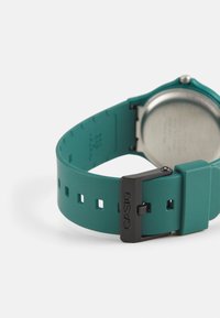 Hodinky Casio s tyrkysovým silikonovým páskem, černým kovovým sponou a stříbrným kovovým zadním víčkem. Páskem jsou vybaveny obdélníkovými výřezy pro nastavení délky.