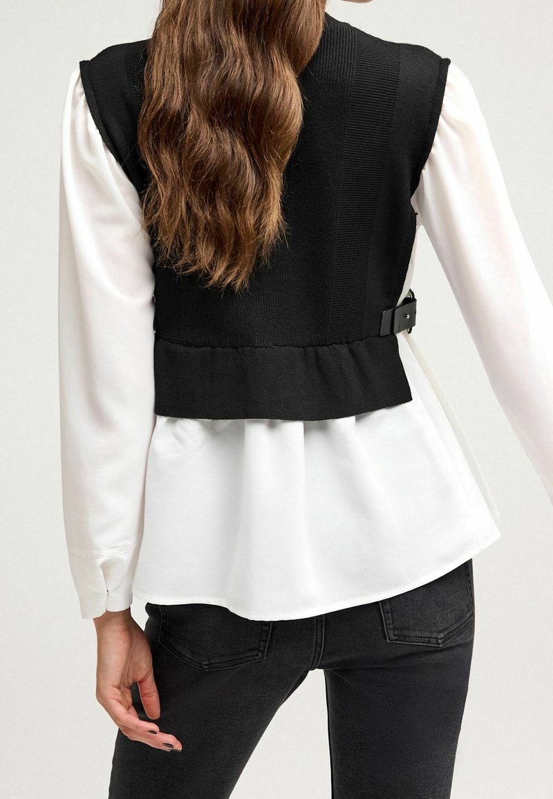 Femme aux longs cheveux bruns portant un chemisier blanc à manches longues et un gilet en maille noir sans manches avec boucle sur le côté, associé à un jean noir.