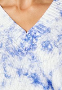 V-ringad tröja i vitt med blå tie-dye-mönster, texturerat stickat material, med ribbade kanter vid halsringningen för extra detalj.