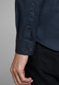 En mans hand bredvid en marinblå texturerad skjortärm med två knappar över en svart byxficka med synlig sömnad.