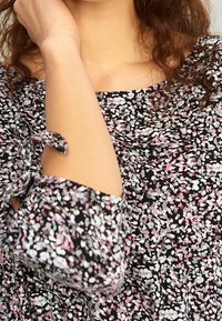 Blusa nera con motivo floreale caratterizzata da un design sparso in rosa e bianco. Scollo curvo con maniche legate e vestibilità ampia. Tessuto leggero.