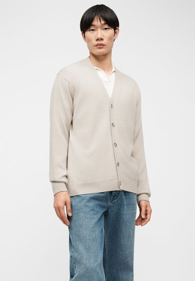 Νέος άνδρας που φοράει μια μπεζ ζακέτα με κουμπιά πάνω από ένα λευκό πουκάμισο και μπλε τζιν, στέκεται μπροστά σε ένα απλό φωτεινό φόντο.