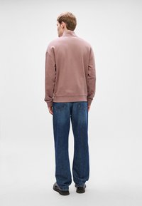 Světle lila mikina s vysokým límcem, žebrovanými manžetami a volným střihem, spojená s volnými tmavě modrými džíny a černými botami.