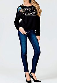 Sweatshirt noir avec broderies florales et texte "CUTE & FIERCE", associé à un jean skinny bleu foncé et des talons noirs.