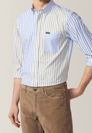 Muž v modré a béžové košili s vertikálními pruhy a náprsní kapsou, s hnědými manšestrovými kalhotami.