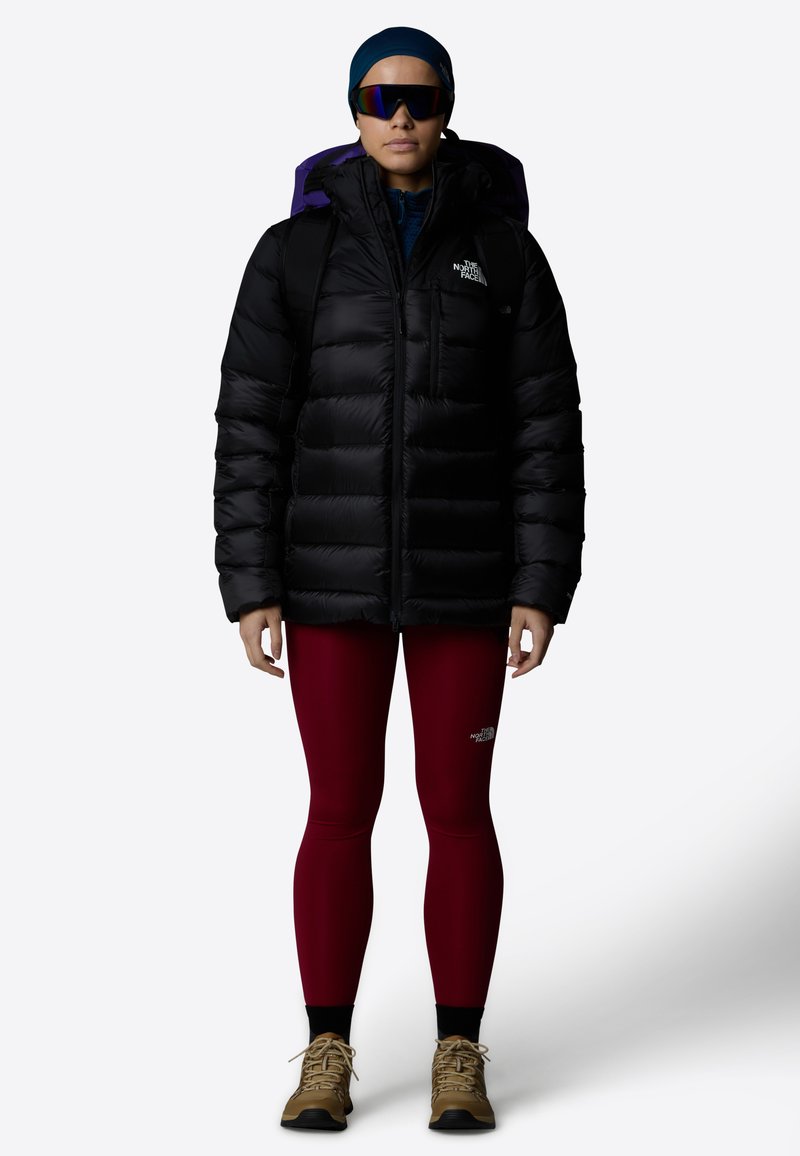 Svart dunjakke med glidelås foran og lilla hette, brukt over marsalalette leggins og brune turbots. Modellen har også solbriller og hodeplagg.