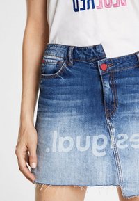 Mujer que lleva una falda de mezclilla azul desgastada con un botón rojo, dobladillo deshilachado y texto impreso en blanco en la parte inferior delantera.