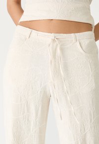 Pantalons en lin blancs avec des motifs floraux brodés, taille élastique et poches avant. Texture douce avec une coupe décontractée.
