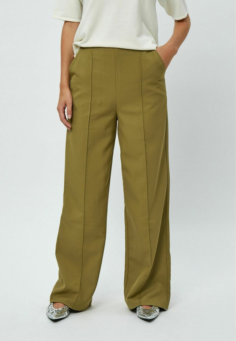 Pantalons en olive vert à jambes larges avec une texture lisse, dotés de poches latérales et de coutures verticales subtiles, associés à des chaussures ornées d'argent.